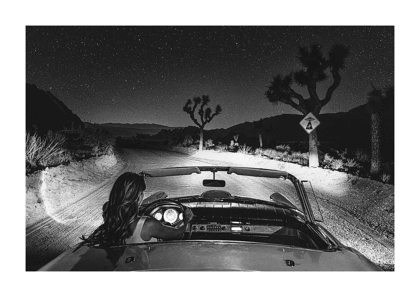 Black and white photograph of a girl driving a vintage convertible down a desert road with a UFO sign warning of alien abductions.