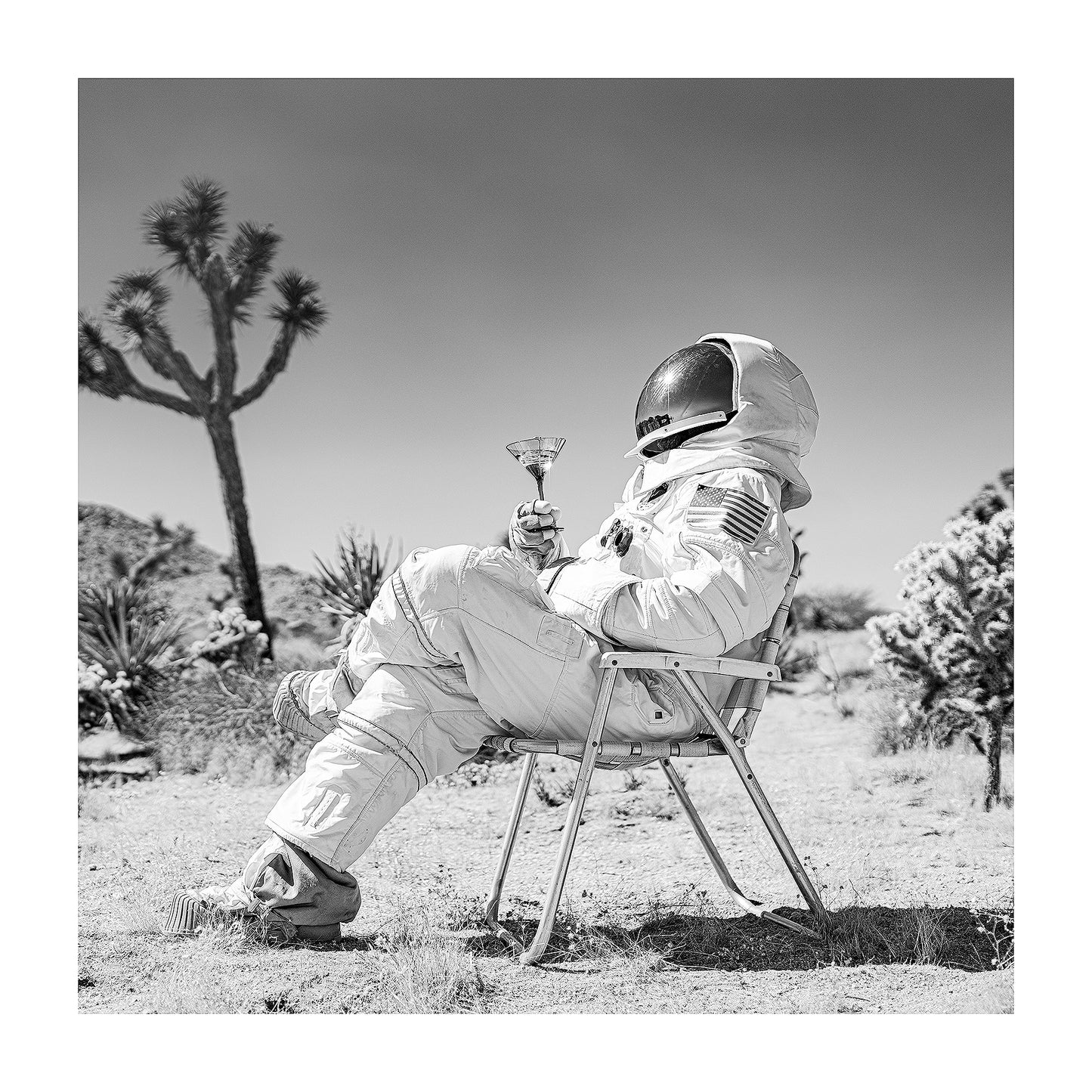 Black and white photograph of an astronaut drinking a martini in a midcentury modern lawn chair in the desert. 