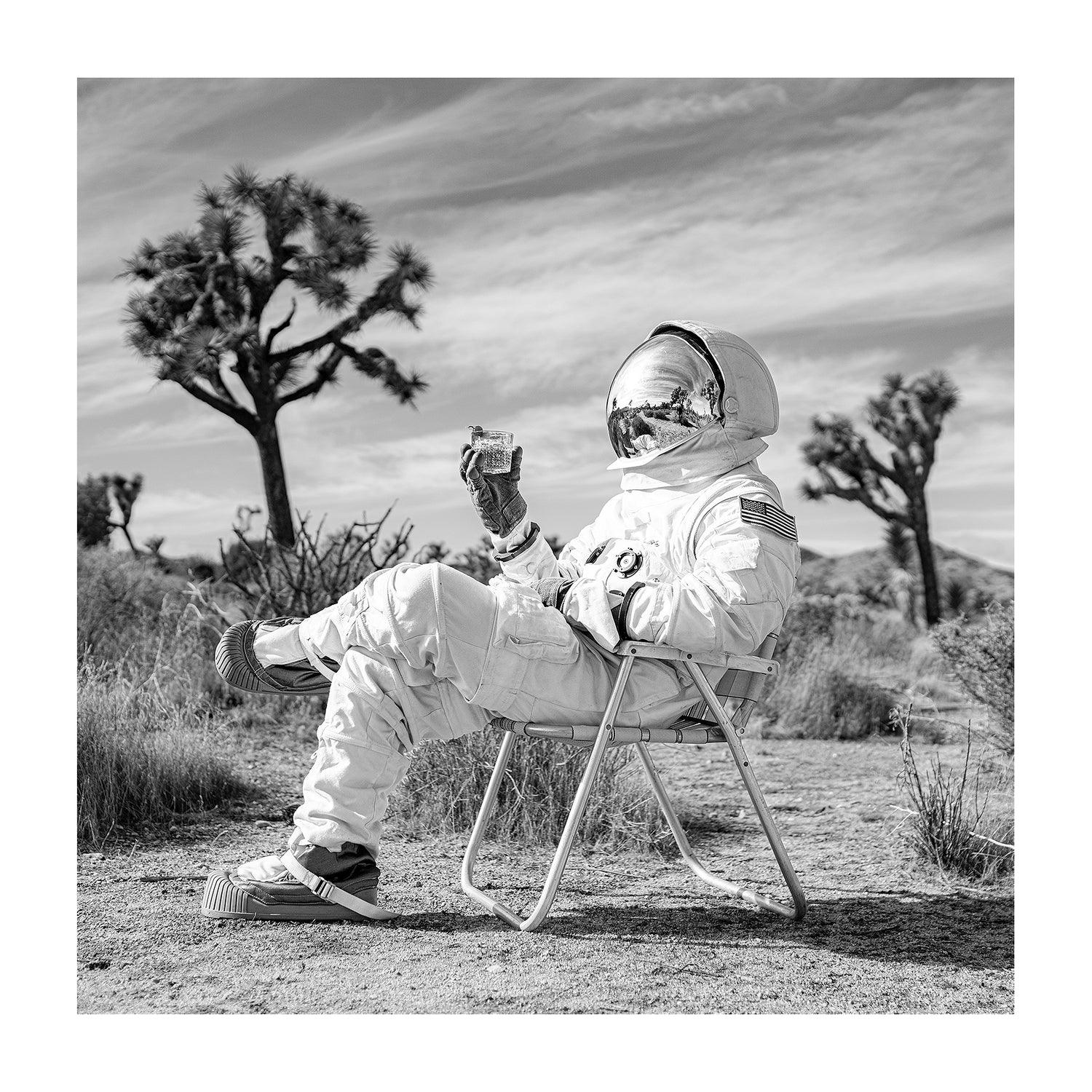 Black and white photograph of astronaut drinking old fashioned cocktail in a midcentury lawn chair. 