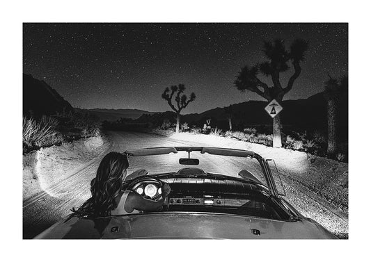 Black and white photograph of a girl driving a vintage convertible down a desert road with a UFO sign warning of alien abductions. 
